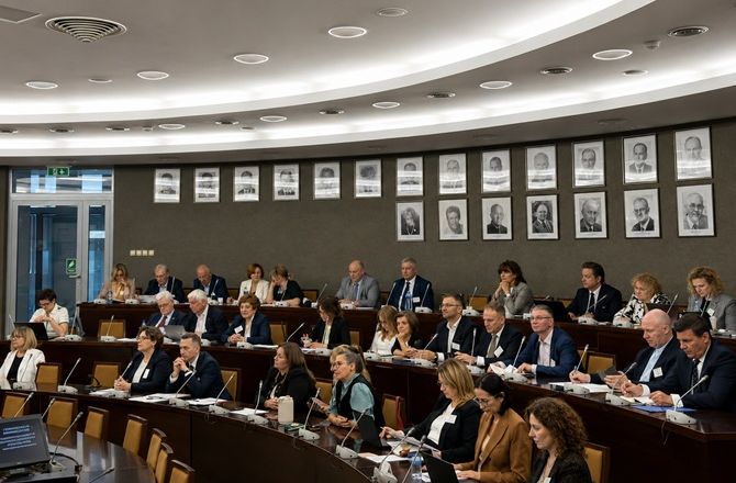 Uczestnicy konferencji siedzą w sali obrad przy wielopoziomowych stołach wyposażonych w mikrofony. Na ścianie widoczne są portrety, a całość ukazuje oficjalny charakter wydarzenia.