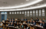 Uczestnicy konferencji siedzą w sali obrad przy wielopoziomowych stołach wyposażonych w mikrofony. Na ścianie widoczne są portrety, a całość ukazuje oficjalny charakter wydarzenia.