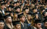 Uśmiechnięte absolwentki i absolwenci w czarnych togach i biretach siedzą wśród uczestników uroczystości.