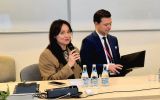 Kobieta i mężczyzna siedzący za stołem prezydialnym. Kobieta przemawia, trzymając w ręku mikrofon.