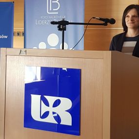 Zbliżenie na postać kobiety za mównicą. Z przodu logo Uniwersytetu Rzeszowskiego. Za mównicą doradca zawodowy Sekcji Biura Karier – mgr inż. Marta Kot. W tle widok posterów informacyjnych Instytutu Ekonomii i Finansów.