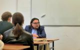 Prelegent siedzi przy stole i odpowiada na pytania studentów podczas spotkania w sali dydaktycznej.