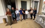 Grupa dziewięciu osób stoi na zadaszonym tarasie zabytkowego budynku, obok pamiątkowej tablicy umieszczonej na ścianie.