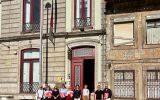 Grupa dziewięciu osób stoi przed wejściem do zabytkowego budynku z czerwonymi drzwiami i ozdobną kamienną elewacją. Część uczestników trzyma czerwone teczki.