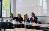 Uczestnicy panelu siedzą przy stole konferencyjnym w sali dydaktycznej. W tle widoczny jest ekran projekcyjny, tablica oraz okna z żaluzjami.