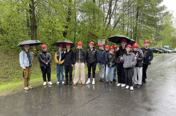 Grupa studentów stojących na zewnątrz, pada deszcz,  w rękach trzymają parasole
