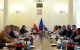 Spotkanie przy długim stole konferencyjnym w reprezentacyjnej sali z flagami Polski i Unii Europejskiej oraz godłem Polski na ścianie.