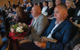 Goście uroczystości siedzą na widowni; jeden z mężczyzn trzyma bukiet kwiatów.