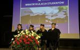 Cztery osoby w strojach akademickich siedzą przy stole prezydialnym na tle prezentacji dotyczącej immatrykulacji studentów.