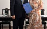 Mężczyzna w garniturze i kobieta w jasnej wzorzystej sukience pozują z granatową teczką w sali posiedzeń.