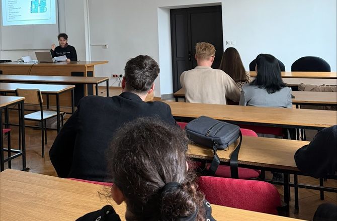 Widok sali wykładowej z uczestnikami siedzącymi w ławkach. Z przodu sali prelegent prowadzi prezentację wyświetlaną na ekranie.