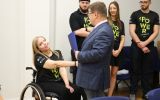 Zawodniczka na wózku inwalidzkim otrzymuje list gratulacyjny od rektora UR. Z tyłu trenerzy oraz inni sportowcy ubrani w klubowe, czarne koszulki z żółtymi napisami "POWER"