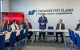 Uczestnicy spotkania siedzą przy stole konferencyjnym na Wydziale Prawa i Administracji Uniwersytetu Śląskiego. Jeden z mężczyzn stoi i przemawia, a w tle widoczny jest ekran z napisem „Konferencja Dziekanów Publicznych Uniwersyteckich Wydziałów Prawa”.