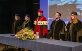Przedstawiciele władz uczelni w strojach akademickich stoją za stołem podczas uroczystej promocji.