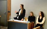 Dwie uczestniczki siedzą przy stole, a jedna stoi i przemawia. Na stole znajdują się dokumenty, książki oraz butelki z wodą.