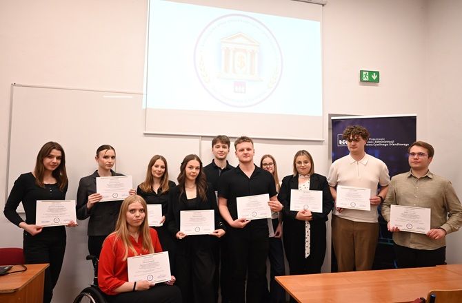 Grupa uczestników seminarium naukowego pozuje do zdjęcia z certyfikatami w sali dydaktycznej. W tle znajduje się ekran projekcyjny.
