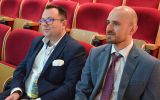 Dwóch uczestników konferencji siedzi w audytorium i obserwuje przebieg obrad.