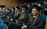 Absolwentki i absolwenci w czarnych togach i biretach siedzą w wypełnionej auli podczas uroczystej promocji.