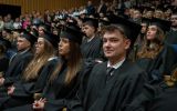 Absolwentki i absolwenci w czarnych togach i biretach siedzą w wypełnionej auli podczas uroczystej promocji.