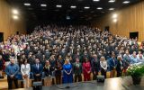 Uczestnicy uroczystości stoją w wypełnionej auli; w pierwszych rzędach znajdują się przedstawiciele władz i zaproszeni goście.