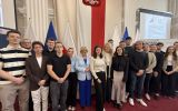 Grupa studentów i uczestniczek debaty „Europa jest kobietą!” pozuje do wspólnego zdjęcia w sali z flagami Polski i Unii Europejskiej.