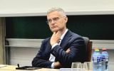 Uczestnik konferencji siedzi za stołem prezydialnym. Przed nim znajdują się materiały konferencyjne, szklanki i butelki z wodą.