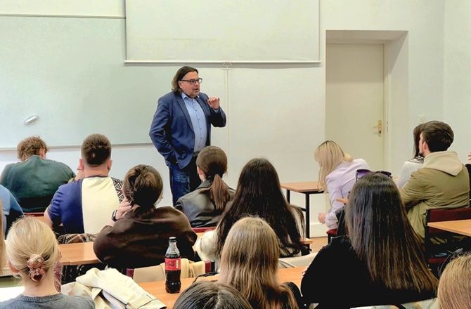 Prelegent przemawia do studentów siedzących w sali dydaktycznej. W tle widoczna jest tablica i ekran projekcyjny.