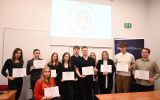 Grupa uczestników seminarium naukowego pozuje do zdjęcia z certyfikatami w sali dydaktycznej. W tle znajduje się ekran projekcyjny.