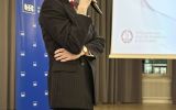 Prelegent w ciemnym garniturze przemawia do mikrofonu obok ekranu konferencyjnego.