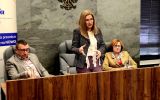 Dwie osoby siedzą przy stole prezydialnym, a jedna osoba stoi obok i przemawia. W tle widoczne jest godło oraz roll-up ELSA Rzeszów.