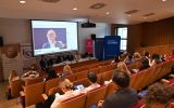 Panel dyskusyjny w sali konferencyjnej z uczestnikami na miejscu oraz prelegentem łączącym się online.