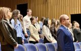 Uczestnicy uroczystości immatrykulacji stoją w rzędach pomiędzy niebieskimi fotelami auli.