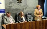 Dwie osoby siedzą przy stole prezydialnym, a jedna osoba stoi obok i przemawia. W tle widoczne jest godło oraz roll-up ELSA Rzeszów.