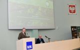 Przedstawiciel lasów państwowych wita uczestników stojąc na mównicy, po prawej siedzi prof. Dudek