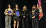 Dwie studentki trzymające niebieskie teczki i czerwone róże pozują na scenie z dwiema przedstawicielkami władz wydziału w strojach akademickich.