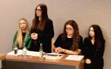 Trzy uczestniczki siedzą przy stole, a jedna stoi i przemawia. Na stole znajdują się dokumenty, książki oraz butelki z wodą.