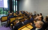 Sala wykładowa z rzędami drewnianych ław, w której zgromadziła się grupa osób biorących udział w konferencji.