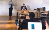 Osoba prowadząca odczytuje materiał przy stole prezydialnym podczas spotkania ze studentami w sali wykładowej.