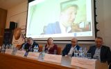 Panel konferencyjny z udziałem prelegenta łączącego się online, widocznego na dużym ekranie.