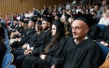 Absolwentki i absolwenci w czarnych togach i biretach siedzą w wypełnionej auli podczas uroczystej promocji.