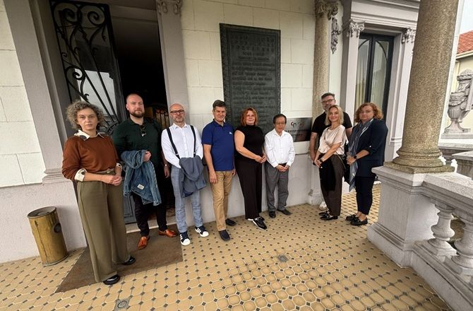 Grupa dziewięciu osób stoi na zadaszonym tarasie zabytkowego budynku, obok pamiątkowej tablicy umieszczonej na ścianie.