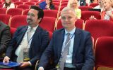 Dwóch uczestników konferencji siedzących w czerwonych fotelach audytorium podczas obrad.