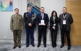 Pięć osób pozuje do oficjalnego zdjęcia w gabinecie; jedna z osób trzyma pamiątkowy dokument.