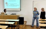 Prowadząca siedzi przy laptopie, a dwie kobiety stoją przy tablicy podczas rozpoczęcia zajęć o kwalifikacjach i predyspozycjach zawodowych mediatora.