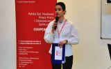 Prowadząca II Forum Cooperante przemawia do uczestników przy banerze wydarzenia.