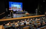 Prorektor przemawia z mównicy podczas uroczystej promocji absolwentów, a przedstawiciele władz uczelni siedzą przy stole na scenie.