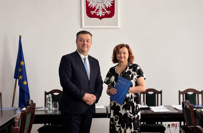 Mężczyzna w garniturze i kobieta w czarno-białej sukience pozują z granatową teczką na tle sali posiedzeń.