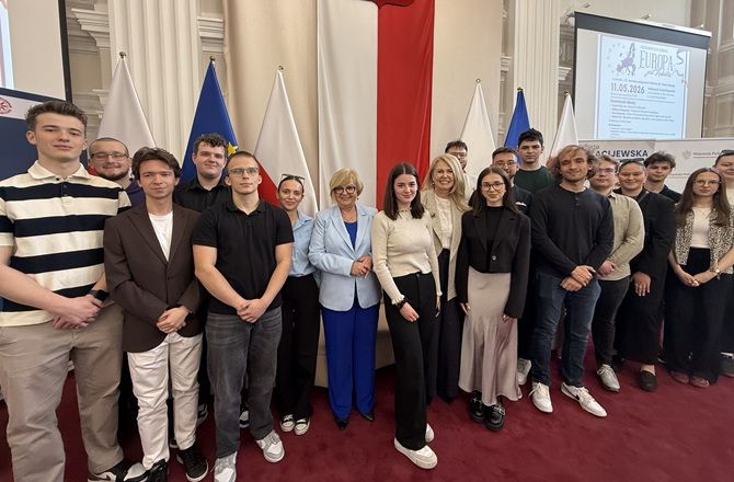 Grupa studentów i uczestniczek debaty „Europa jest kobietą!” pozuje do wspólnego zdjęcia w sali z flagami Polski i Unii Europejskiej.
