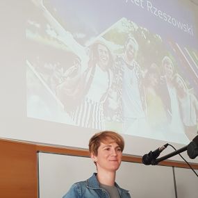 Zbliżenie na postać kobiety za mównicą. Z przodu logo Uniwersytetu Rzeszowskiego. Za mównicą pracownica Sekcji Rekrutacji – mgr Karolina Mróz, która omawia przebieg procesu rekrutacyjnego. W tle slajd, na którym widać napis „Uniwersytet Rzeszowski”