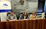 Trzy osoby siedzące przy stole prezydialnym. W tle widoczne jest godło oraz roll-up ELSA Rzeszów.